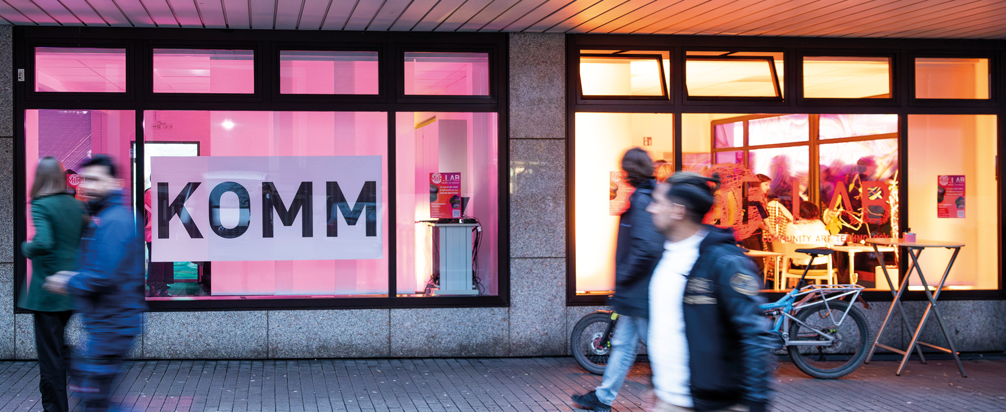 Ein Schaufenster, welches mit rosa Licht beleuchtet wird und eines mit gelbem Licht. Im rosa Fenster steht in großen Buchstaben "KOMM". Menschen Laufen vorbei und schauen hinein.