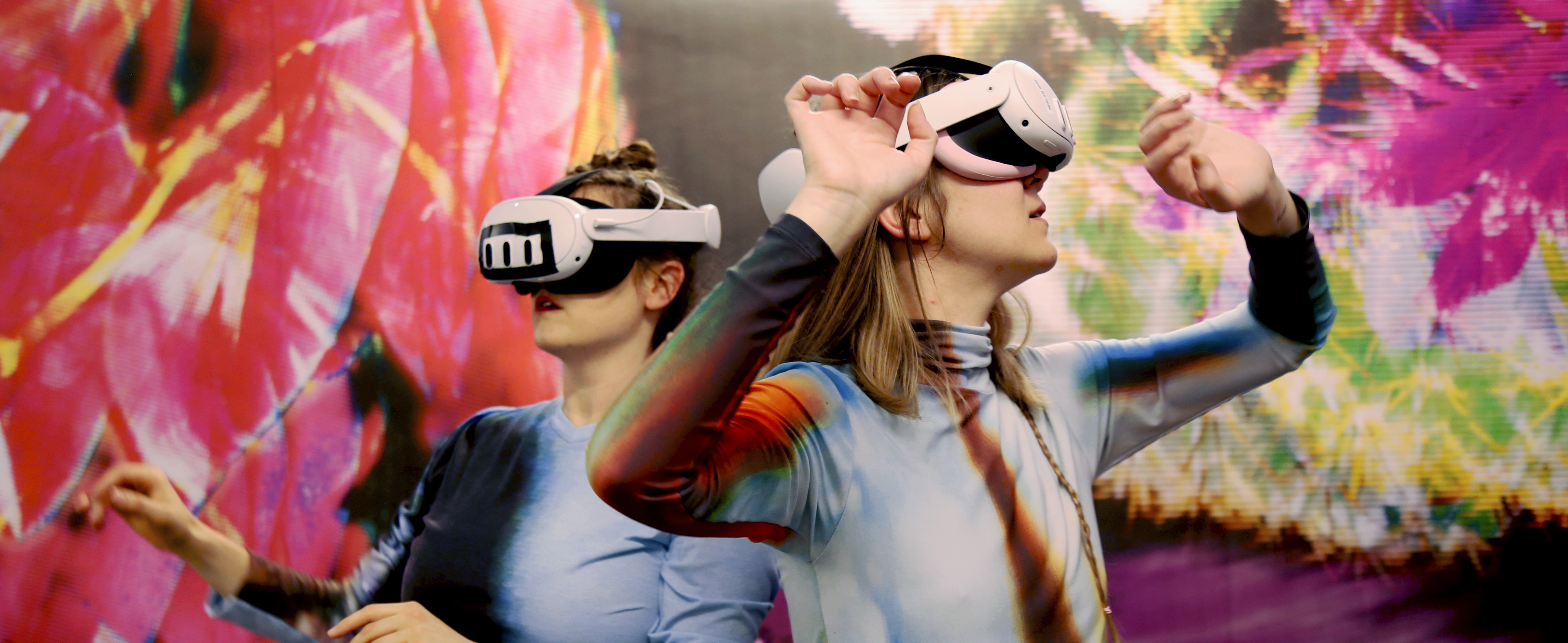 Zwei Personen, welche sich mit VR-Brillen umsehen. Im Hintergrund bunte Struktur.