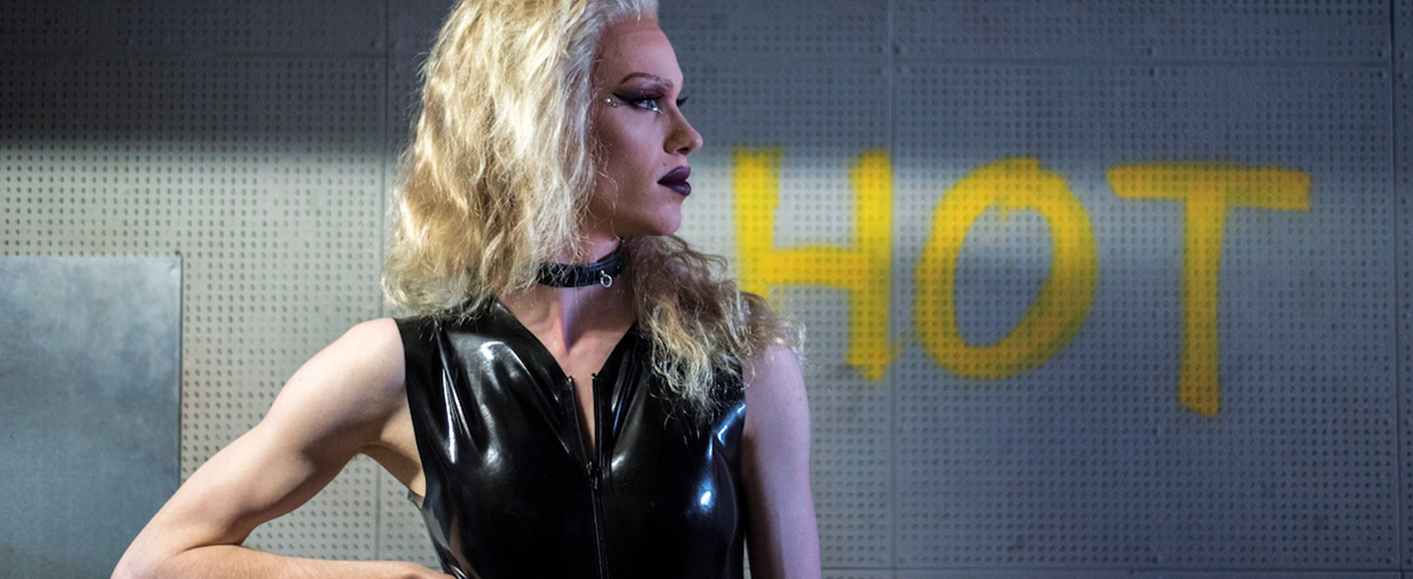 Eine Dragqueen mit blonder Perücke und schwarzem Lackoberteil im Profil mit blick nach rechts. Im Hintergrund steht in gelber Schrift auf der Wand das Wort "HOT"