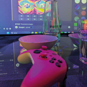 Ein pinker Controller auf einem Glastisch neben einem Glas, zwei Schüsseln und einer Glasflasche. im Hintergrund eine Projektion
