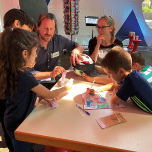 Vier Kinder und zwei Erwachsene sitzen an einem Tisch und schreiben auf Zettel. Auf dem Tisch steht ein Fußball mit einer Schirmmütze.