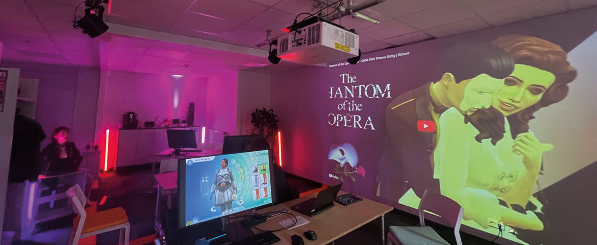 Ein Raum voll mit Computertechnik, leuchtenden Bildschirmen und Leuchtröhren. An der Wand die Projektion von zwei cartoonartigen Personen, welche sich in der Armen liegen. Daneben der Schriftzug "The Phantom of the Opera".