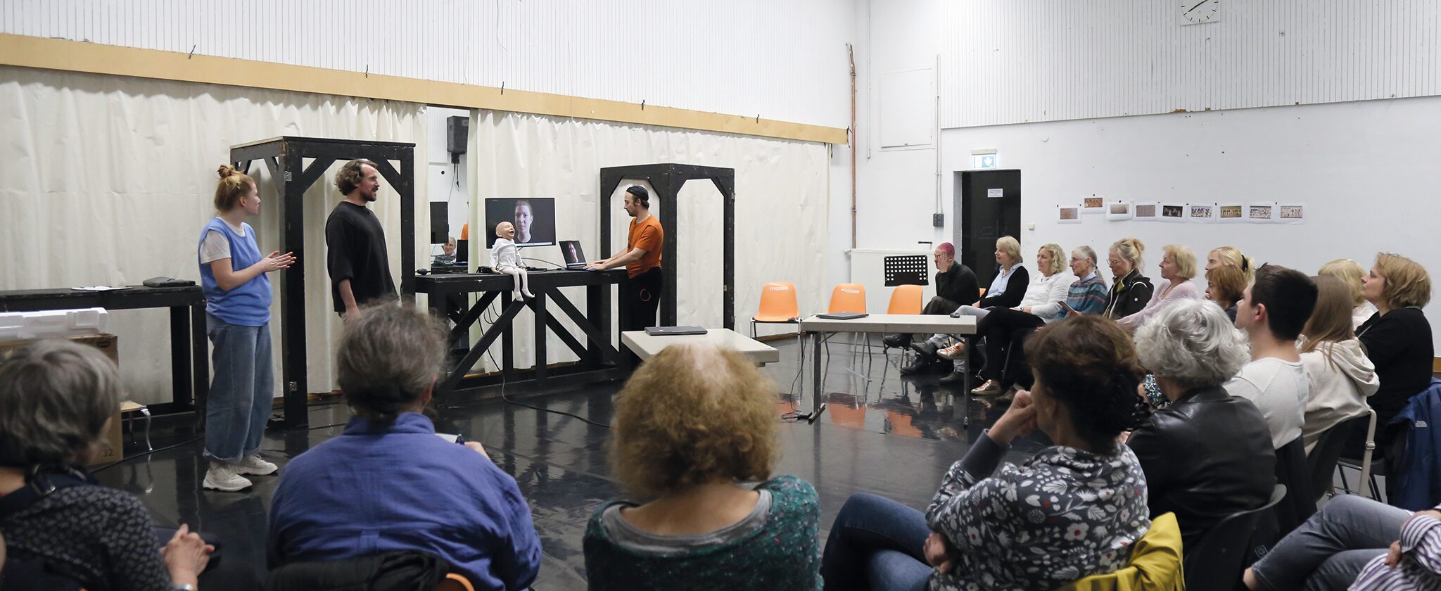 Viele Menschen sitzen im Halbkreis, während vorn drei Personen referieren. Auf einem Tisch sitzt eine Puppe neben einem Bildschirm, welcher einen Avatar zeigt.