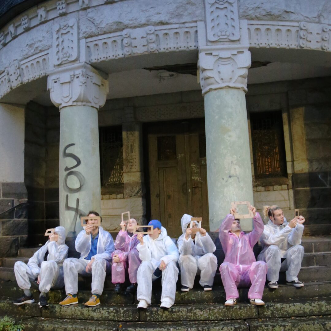 Sieben Personen sitzen in Maleranzügen vor dem Eingang einer Kirche und gucken durch rechteckige Papierrahmen.