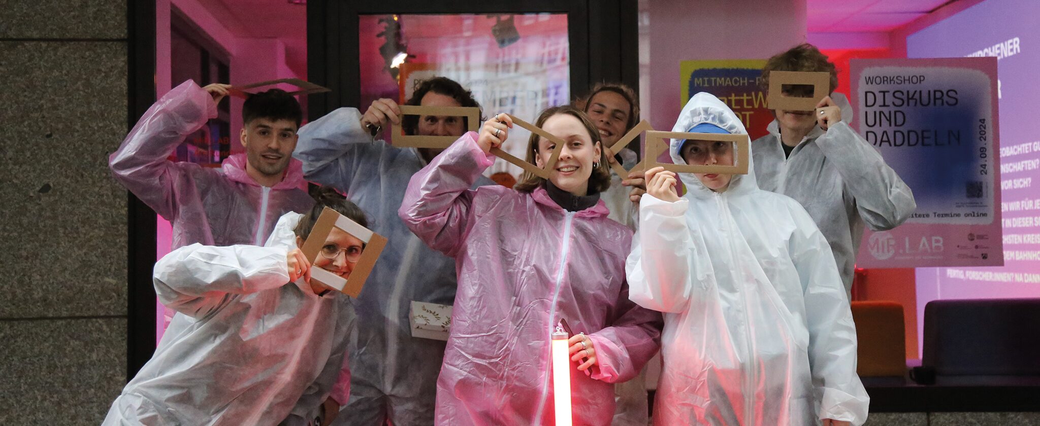 Sieben Personen posen in Maleranzügen mit rechteckigen Papierrahmen und einer LED-Röhre vor dem MiR.LAB.