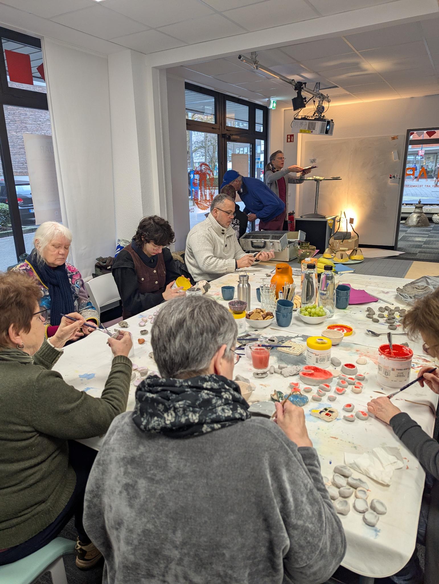 Eine Gruppe Menschen sitzt an einem Tisch und bemalt kleine Tonskulpturen.