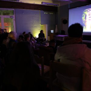 Menschen schauen auf einen großen Bildschirm, wo man eine Frau mit geschlossenen Augen sieht.