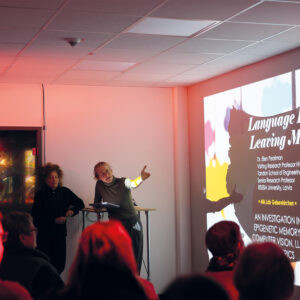 Mehrere Menschen sitzen vor einer Projektion. Neben deben dem projizierten Bild stehen zwei Personen und erläurtern.