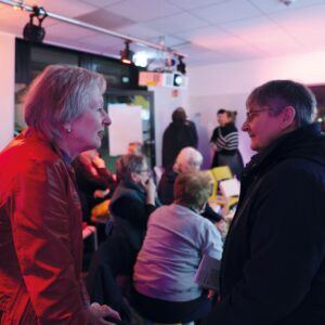 Im Vordergrund zwei Personen tief in ein Gespräch vertieft. Im Hintergrund weitere Menschen auf Stühlen sitzend