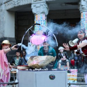 Drei Personen in aufwändigen Kostümen beschwören eine merkwürdige Skulptur. Die Person links bedient eine kleine Nebelmaschine, die Person rechts spricht in ein Megafon.