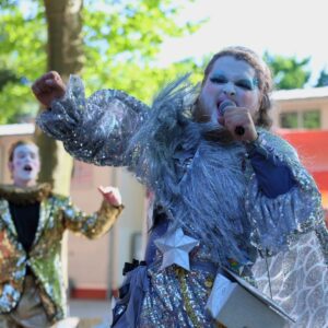 Eine Person mit Bart, Make-Up und silbernem Kostüm singt enthusiastisch in ein Mikrofon. Im Hintergrund verschwommen ein König im goldenen Kostüm.