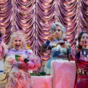 Drei Frauen in fantasievollen, barock anmutenden Kostümen mit weißen Perücken essen farbige Gelees an einem dekorativen Tisch vor einem rosa-glänzenden Vorhang. Die Szene wirkt theatralisch und übertrieben inszeniert.