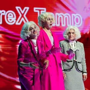 Drei Personen mit blonden Perücken und eleganter Kleidung stehen vor einem leuchtend roten Hintergrund mit Schriftzug. Zwei tragen pinke Kostüme, die dritte ein schwarz-weißes Hahnentrittmuster – eine stilisierte Darstellung älterer Damen.