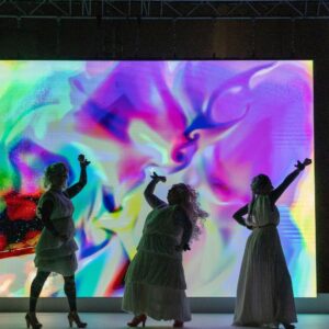 Drei Frauen in weißen Gewändern und mit erhobenen Armen stehen silhouettenhaft vor einem bunten, abstrakten LED-Hintergrund. Die Szene wirkt tänzerisch und feierlich.