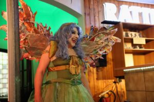 Eine Person im grünen Kostüm mit lila Perücke und Flügeln aus Durstlöschern lacht herzlich.