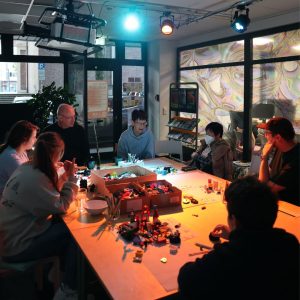 In dem Bild ist in der Mitte ein großer Tisch zu erkennen auf dem Boxen mit Legosteinen stehen. Um den Tisch herum sitzen Personen.