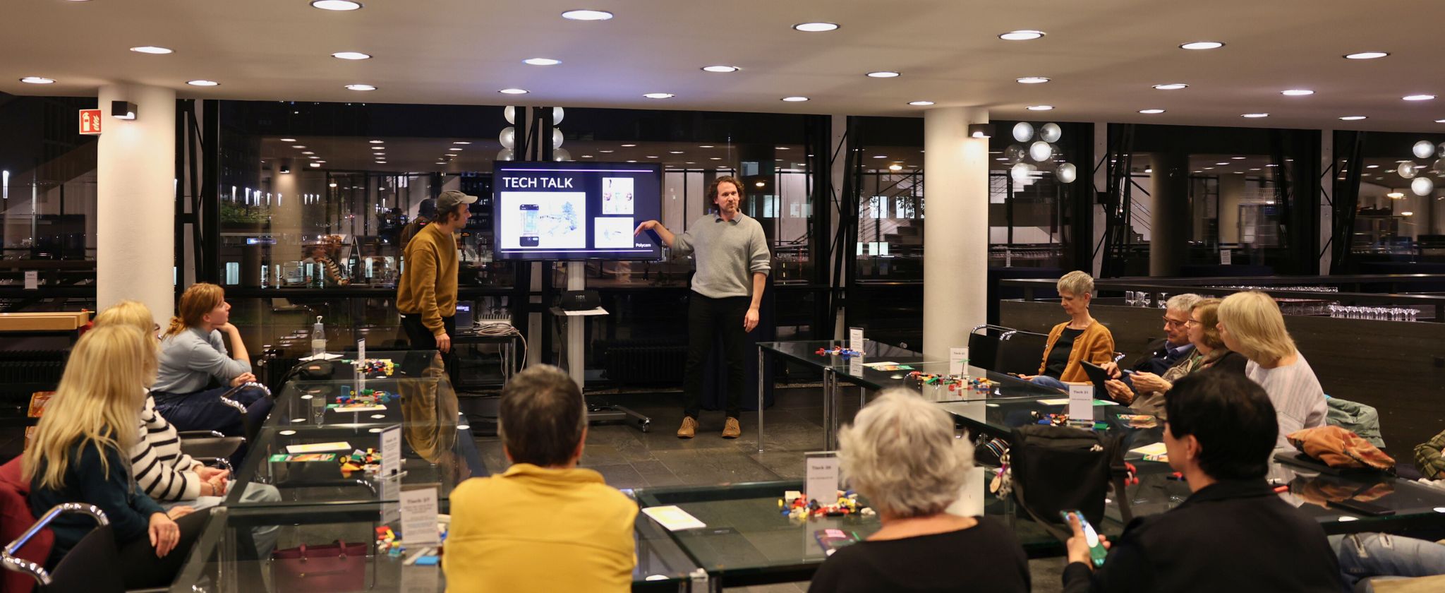 Dominik Hallerbach erklärt vorne an einem Bildschirm etwas über die Software ,,Polycam'' und Tech Talk. Vor ihm sitzen Menschen mit Lego und Ipads auf Ihren Tischen, für 3D Scans.