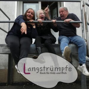 Gruppenfoto der Langstrümpfe. Sie sitzen auf einer Art Podest und halten sich an einem Geländer fest. Im Vordergrund steht in einer Blase: "Langstrümpfe - Rock für Kids und dich".