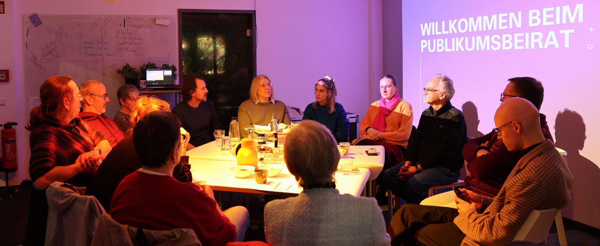 In dem Bild sieht man einen Tisch in der Mitte um den ganz viele Menschen herum sitzen.