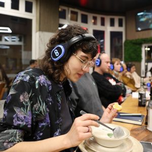 Im Bild sieht man eine Junge Frau sitzen am Tisch. Sie trägt Kopfhörer und ist dabei eine Suppe.