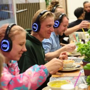 Im Bild sitzen Personen mit Kopfhörern am Tisch und Essen Suppe.