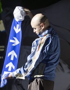Parent hält blauen Schal mit Pfeilen in der Hand. Er tragt Kappe und Jacke schaut nach unten dunkler Bühnenhintergrund.