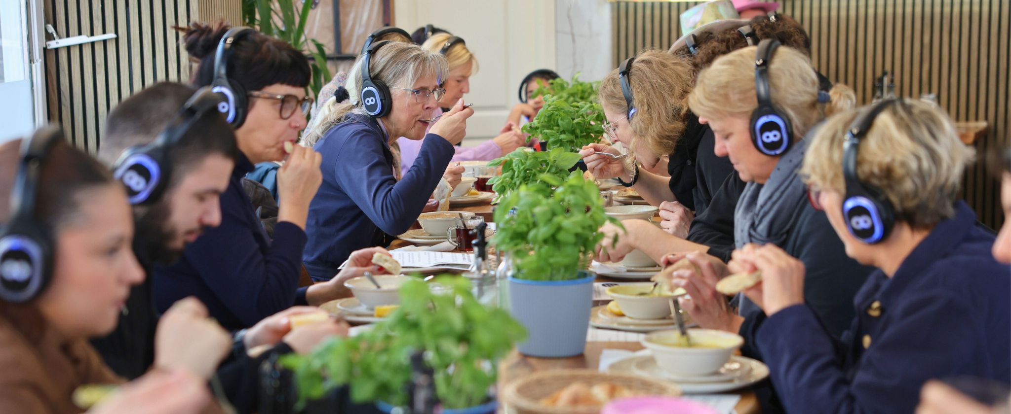 In der Mitte von Bild befindet sich ein langer Tisch mit Essen drauf. Rechts und Links vom Tisch sitzen Menschen und Essen Suppe.