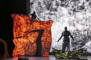 Teen steht oben links auf Kissenkulisse, Ghost unten rechts. Kulisse rot beleuchtet, Hintergrund hell, Bühne bedrohlich.