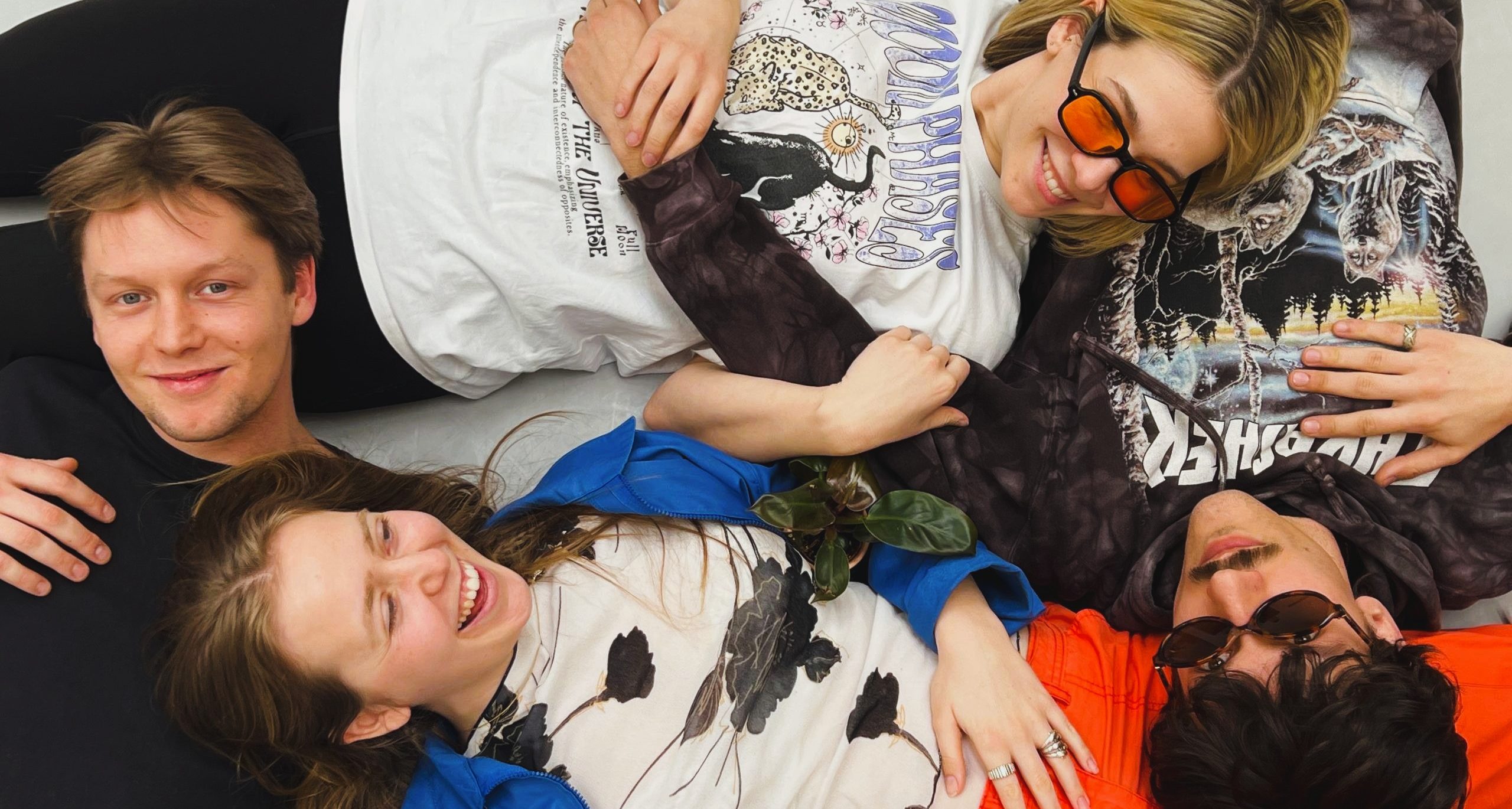 Die Küsntler*innen Sandro, Annika, Victoria und Daniel liegen im Kreis auf dem Boden. Ihre Köpfe liegen jeweils auf den Bäuchen oder Beinen der anderen. Sie tragen lockere Kleidung und zwei eine Sonnenbrille. Sie lachen. Sie werden aus der Vogelperspektive fotografiert.