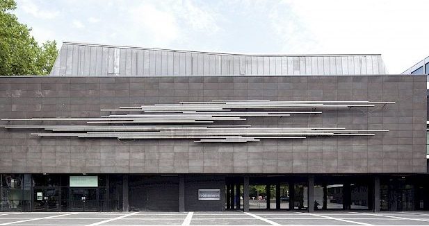 Das Gebäude vom Kleinen Haus von außen fotografiert. Blauer Himmel. Bäume an der linken Seite. Großer Platz davor. Graue Fassade.