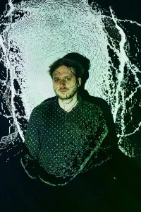 Portraitfoto von Dan Molnar. Braune Haare, dunkles Hemd, steht in Projektion