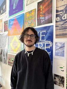 Portraitfoto Darius. Braune Haare, Brille, Bart, vor Wand mit Plakaten