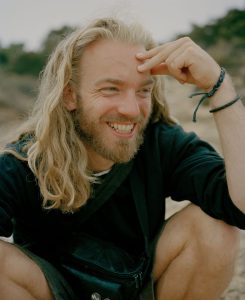 Portraitfoto Felix, lange blonde Haare, Bart, sitzt, lacht, am Strand