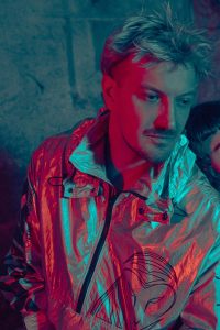 Portraitfoto Lex, kurze blonde Haare, Bart, glänzende Jacke, rot grünes Licht