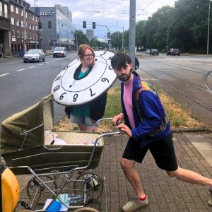 Eine Person mit Uhren Kostüm und die andere mit einem Kinderwagen auf der Straße.