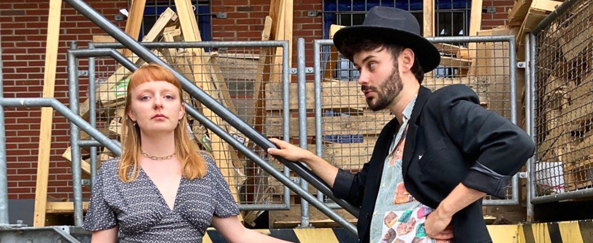 Maj und Anton posieren auf einer Metalltreppe vor einer Backsteinwand mit gestapelten Holzpaletten. Maj steht links in einem gemusterten Jumpsuit und schwarzen Absatzschuhen, Anton rechts in Anzug, Hut und auffälligen orangefarbenen Socken, mit einem Fuß auf der Treppenstufe.